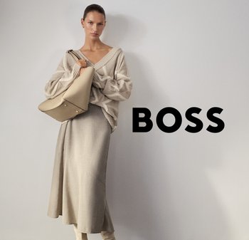 Vrouw in een beige v-hals trui en een lange rok, met een grote beige handtas, staand naast zwarte tekst "BOSS" op een grijze achtergrond.