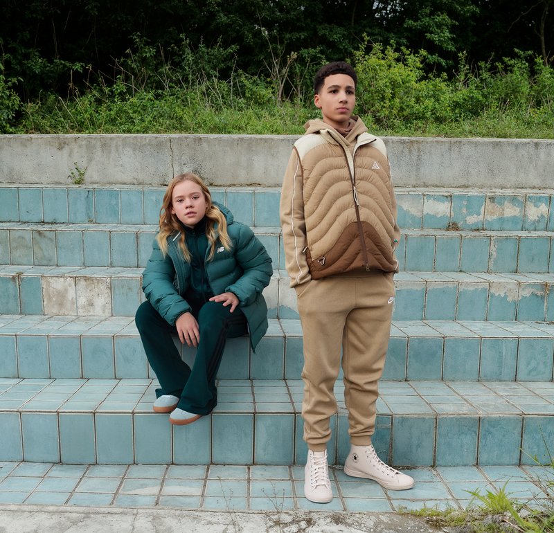 Zwei Personen sitzen auf einer blau gefliesten Treppe. Die eine trägt eine türkisfarbene, isolierte Jacke und passende Hosen; die andere trägt eine beige gesteppte Jacke und beige Hosen.