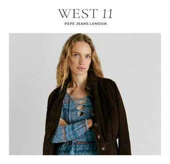 Woman with wavy blonde hair wearing a blue lace-up knit top under a dark brown jacket, posing with arms crossed against a plain background.