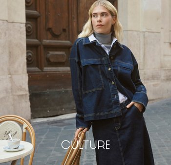 Blonde Frau in dunkler Jeansjacke und Rock steht auf einer Kopfsteinpflasterstraße und lehnt ihre Hand an einen Holzstuhl neben einem Café-Tisch.