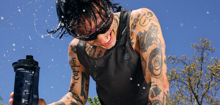 Personne tatouée portant des lunettes de soleil et un débardeur verse de l'eau sur elle-même à l'extérieur, tenant une grande bouteille d'eau, avec un ciel bleu et des arbres en arrière-plan.