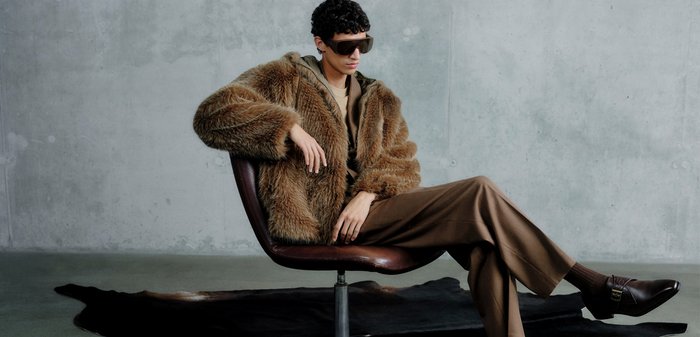 Manteau en fausse fourrure marron à coupe ample, assis sur une chaise en cuir marron, associé à un pantalon marron et des chaussures en cuir foncé, fond gris.