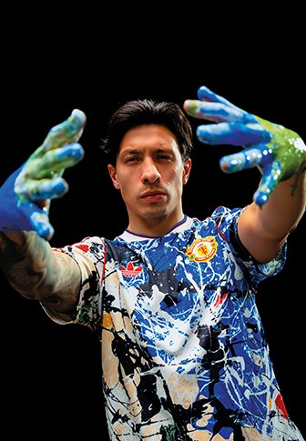 Man met verf bedekte handen die naar voren reikt, gekleed in een kleurrijk gespat shirt tegen een zwarte achtergrond.