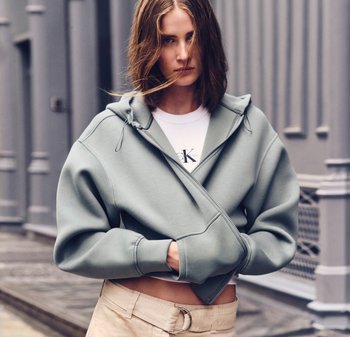 Ung kvinna med axellångt hår som bär en blekgrön oversized zip-up hoodie och beige byxor, ståendes utomhus vid en grå byggnad.