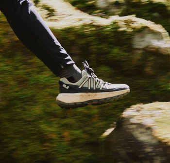 Trailrunning-Schuh in Grau und Schwarz, mit einer strukturierten Sohle, Schnürsenkeln und prominenter Dämpfung, über einem moosbedeckten Boden in Bewegung.
