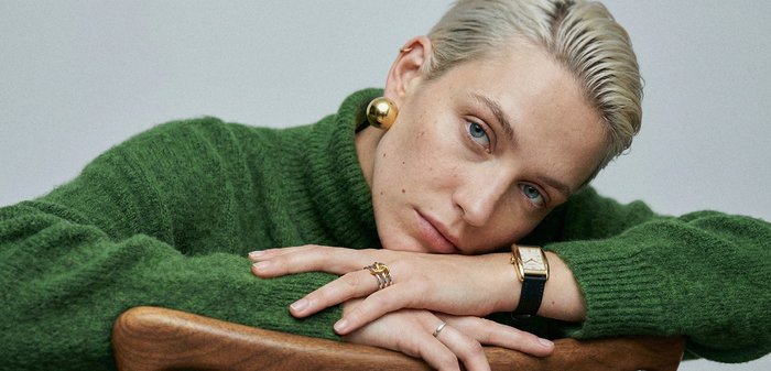 Personne aux cheveux blonds courts reposant les bras sur une chaise en bois, portant un pull vert, des boucles d'oreilles en or, des bagues et une montre-bracelet.