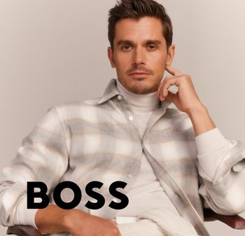 Uomo con capelli corti castani che indossa una camicia a quadri beige e un dolcevita crema, seduto con la mano vicino al viso; testo nero "BOSS" nell'angolo in basso a sinistra.