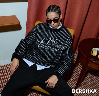 Černobílý pruhovaný pletený svetr s chlupatým efektem a výrazným nápisem. Modelka nosí sluneční brýle a sedí na židli.