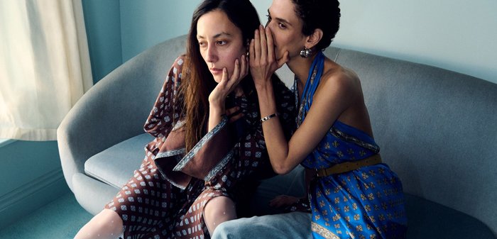 To kvinder sidder; den ene i brun mønstret kjole og hvide hæle ser betænksom ud, den anden i blå mønstret top hvisker i hendes øre.