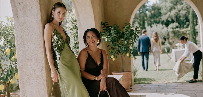 Deux femmes en robes élégantes, l'une debout en vert et l'autre assise en noir, posent sous des arches avec des citronniers, pendant qu'un couple marche en arrière-plan.