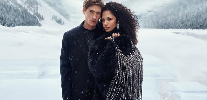 Abrigo de piel negra y bolso adornado con detalles plateados llevados por dos modelos en un paisaje nevado. Copos de nieve caen sobre un fondo de tonos azul hielo.