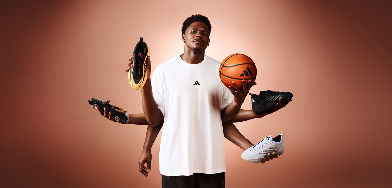 Hombre con camiseta blanca de Adidas sosteniendo un balón de baloncesto y cuatro zapatillas deportivas diferentes frente a un fondo marrón liso.