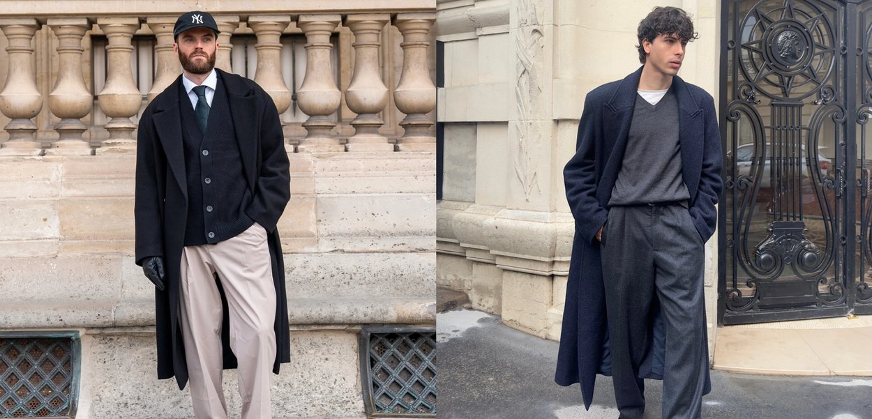 Kaksi miestä poseeraa ulkona kivisten rakennusten edessä; molemmilla on päällään pitkät tummat takit kerrospukeutumisen päällä, ja heillä on rennot housut.