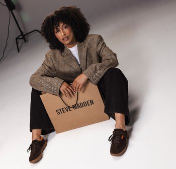Bruine ruitblazer met zilveren knopen, witte t-shirt, zwarte wijde broek en bruine sneakers. Houdt een bruine Steve Madden winkeltas vast.