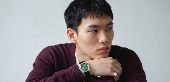 Jeune homme en pull bordeaux reposant le menton sur les bras croisés, portant une montre à cadran vert et des bagues, regardant pensivement sur le côté.