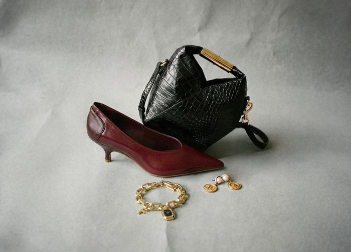 Burgundy leather high-heeled shoe, black croc-embossed handbag, gold bracelet, and earrings featuring pearl and coin designs on a grey background.