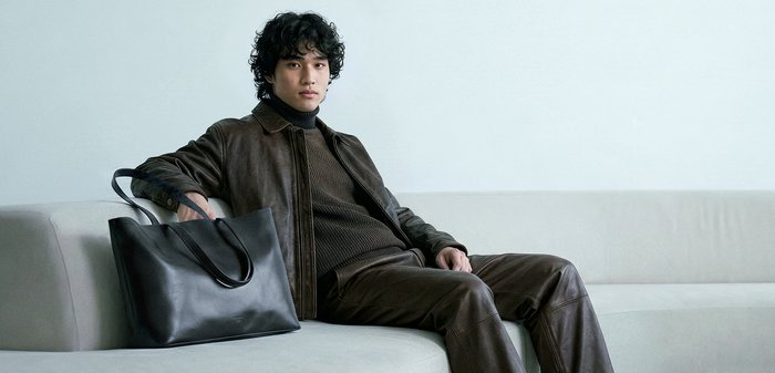 Νέος άνδρας με καφέ μπουφάν και παντελόνι κάθεται σε ανοιχτό γκρι καναπέ με μεγάλη μαύρη δερμάτινη τσάντα ώμου δίπλα του.