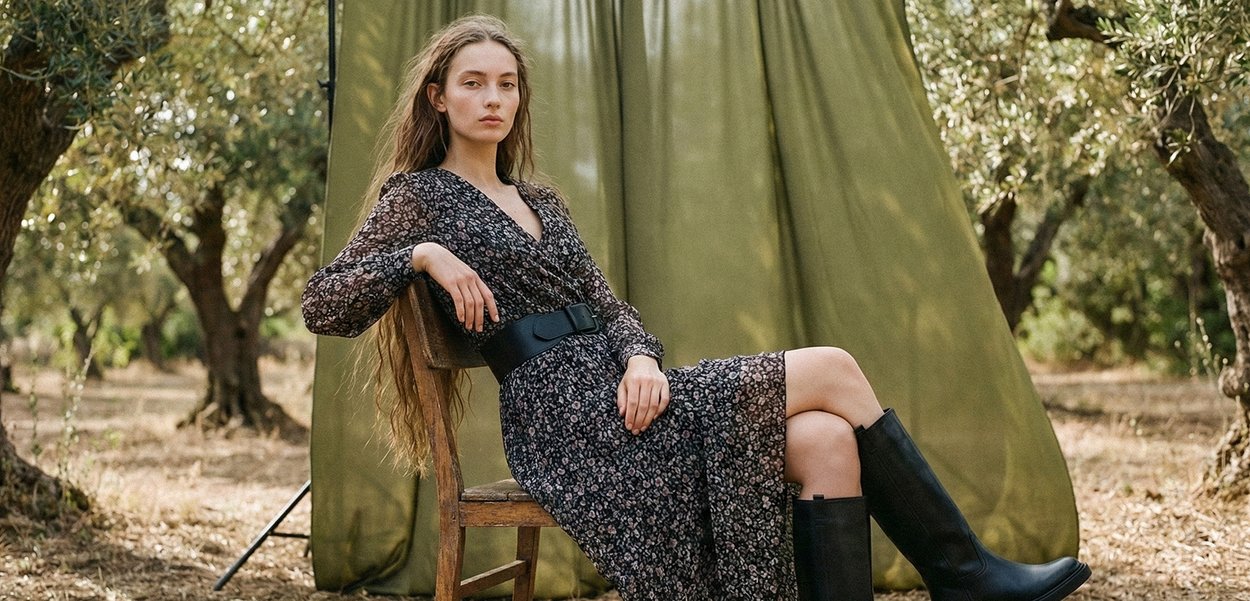 Mujer con cabello largo, vestido estampado y botas negras sentada en una silla de madera al aire libre entre árboles de olivo.