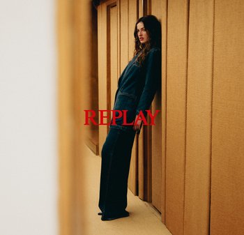 Denim overall i mörkblått, med vida byxor och långa ärmar. Modellen lutar sig mot en texturerad trävägg. Röd "REPLAY"-logotyp syns.