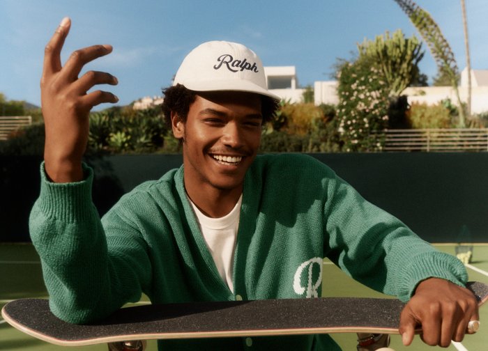 Joven sonriente con gorra blanca de Ralph y cárdigan verde sosteniendo una patineta en una pista de tenis al aire libre a la luz del día.