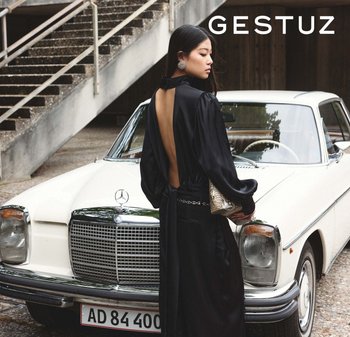 Crna haljina bez leđa s dugim rukavima i vezanjem u struku, uparena s torbicom od zmijske kože, pored vintage bijelog Mercedesa.