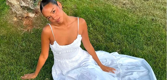 Femme en robe blanche assise sur l'herbe verte près d'un arbre, regardant vers le haut avec une expression détendue.