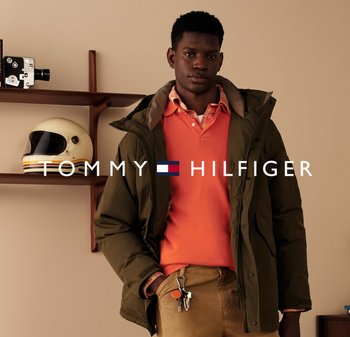 Zielona kurtka puchowa z kapturem, noszona na pomarańczowym polo i koszuli w kratę. Przy pasie widoczny brelok. Proste tło.