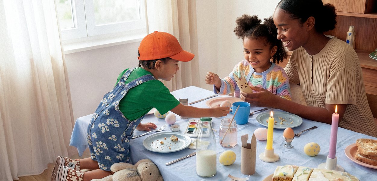 Dos niños y una mujer pintando huevos de Pascua juntos en una mesa con aperitivos, velas y material de arte en una habitación luminosa.