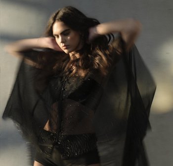 Top nero trasparente con decorazioni, sovrapposto a un body coordinato. I capelli della modella sono ondulati e il tessuto appare traslucido con un movimento fluido.