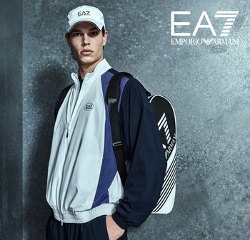 Veste zippée en gris et marine avec logo. Le modèle porte une casquette blanche et porte un sac à dos noir à motif rayé blanc. Texture de tissu lisse.