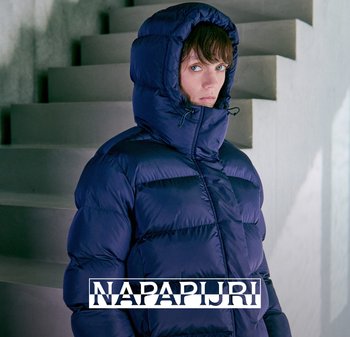 Persona che indossa un giubbotto imbottito blu scuro con il cappuccio alzato, in piedi all'interno vicino a una scala in cemento, che guarda leggermente di lato.