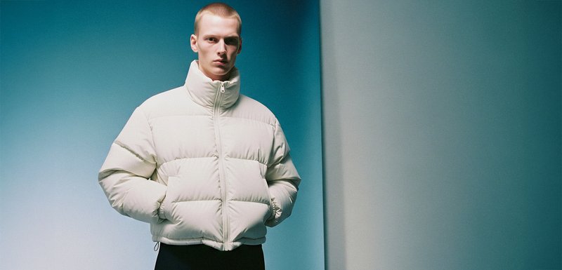 Veste blanche rembourrée avec un col haut, une fermeture éclair et des poches latérales. Tissu texturé visible en panneaux horizontaux.