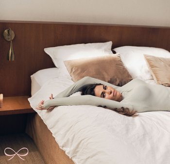 Langarm, lichtgroen top op een persoon die op een witte dekbed ligt met beige kussens. Houten nachttafeltje met een gouden wandlamp.