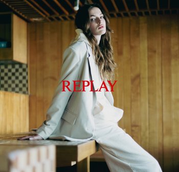 Blazer e pantaloni su misura di colore chiaro in un tessuto morbido, con la parola "REPLAY" in evidenza in rosso. Sfondo in legno.