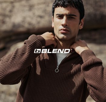 Maglione marrone lavorato a maglia con collo zip e texture a coste. Modello che sistema il colletto, sullo sfondo di rocce. Logo del marchio visibile.