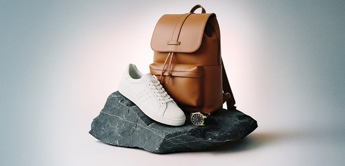 Καφέ δερμάτινο σακίδιο, λευκό αθλητικό παπούτσι και μαύρο ρολόι χειρός με χρυσή επένδυση τοποθετημένα σε σκούρα υφή πέτρας.