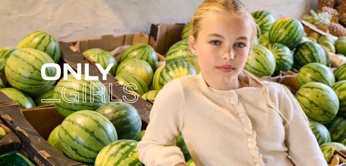 Una niña con cabello largo y rubio viste un cárdigan beige claro con detalles de volantes, de pie junto a una pila de sandías verdes.