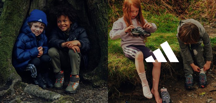 Deux enfants sont assis près d'un arbre, vêtus de vêtements de plein air noirs et gris. Un enfant porte des chaussures sombres avec des accents lumineux ; l'autre ajuste des chaussures vertes sur l'herbe.