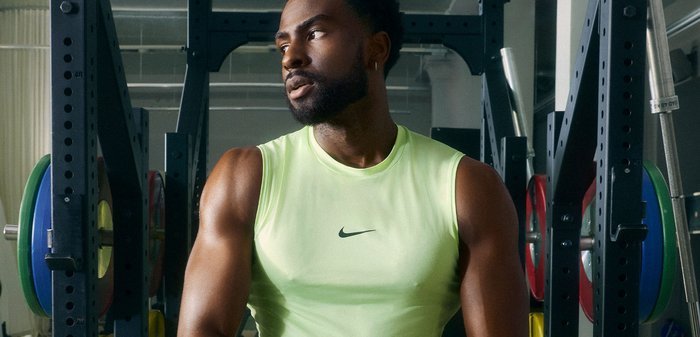 Férfi ujjatlan neon zöld Nike pólóban áll egy edzőteremben, súlyemelő állvány mellett, színes tárcsákkal.