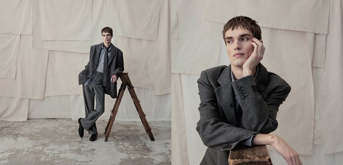Jovem de fato cinzento apoia-se numa escada de madeira contra um fundo de tecido bege texturado, olhando pensativamente para o lado.