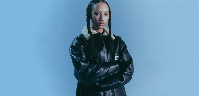 Giacca nera in pelle con colletto in shearling crema, dotata di chiusura frontale con bottoni e tasche laterali. Il modello ha lunghi capelli intrecciati.