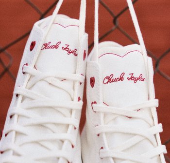 Zapatillas altas blancas con cordones rojos, con "Chuck Taylor" bordado en rojo y ojales en forma de corazón. Textura lisa de lona.