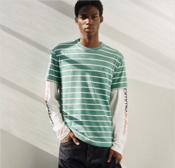 Hombre joven con camiseta de manga corta de rayas verdes y blancas sobre una camiseta blanca de manga larga, de pie en interiores con la mano en el bolsillo.