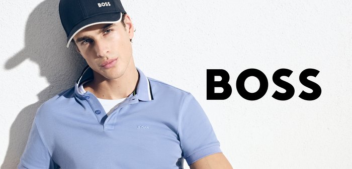 Jonge man in een blauwe polo en zwarte pet met het "BOSS"-logo, staand tegen een witte muur met grote "BOSS"-tekst naast hem.