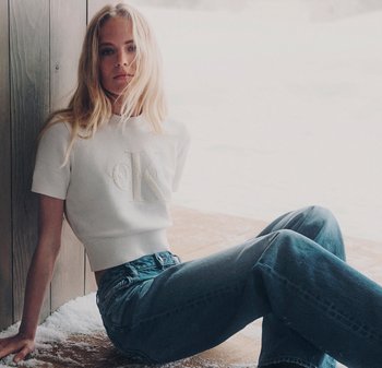 Vitt stickat kort sweatshirt med en texturerad "K"-logotyp och korta ärmar, tillsammans med högmidjade blå denimjeans, sittande i en snöig miljö.