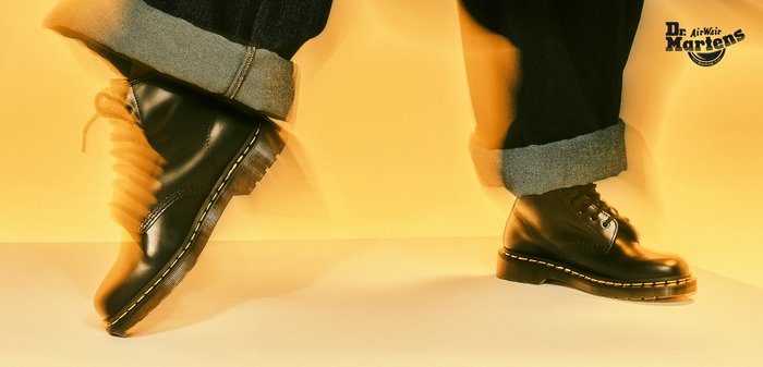 Schwarzer Dr. Martens Stiefel aus Leder mit gelber Naht, schwarzen Schnürsenkeln und gebrandetem Laschen-Label, vor hellem Hintergrund gezeigt.