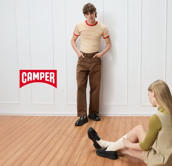 Helle beige T-Shirt mit rotem Saum, braunen weit geschnittenen Hosen und schwarzen Schuhen. Holzboden; eine sitzende Person in beigem Kleid und cremefarbenen Socken.