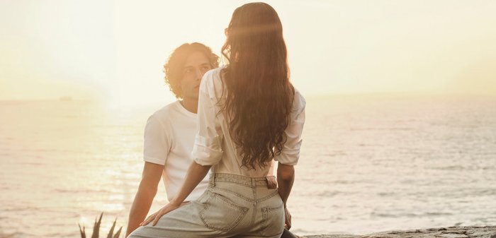 Couple assis au bord de la mer au coucher du soleil, se faisant face avec une lumière douce et un océan calme en arrière-plan.