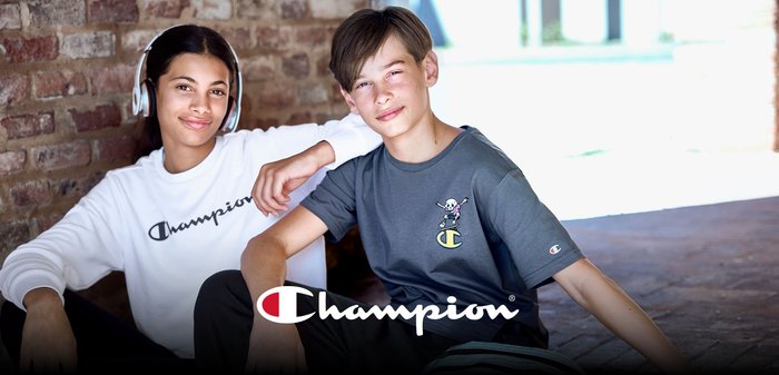 Dois jovens sentam-se encostados a uma parede de tijolos. Um veste uma camisola branca com a palavra "Champion" em letra cursiva, o outro uma camisa cinzenta com um desenho gráfico.