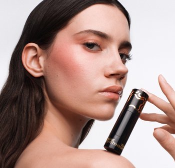 Selkeä ihoinen nainen pitää mustaa ja kultaa olevaa sylinterin muotoista ihonhoito- tai meikkituotetta olkansa lähellä valkoista taustaa vasten.